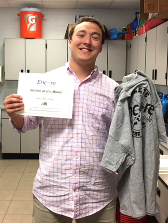 Young man smiling and posing for photo holding a paper certificate and t-shirt which read Encore Sports Medicine Athlete of the Month